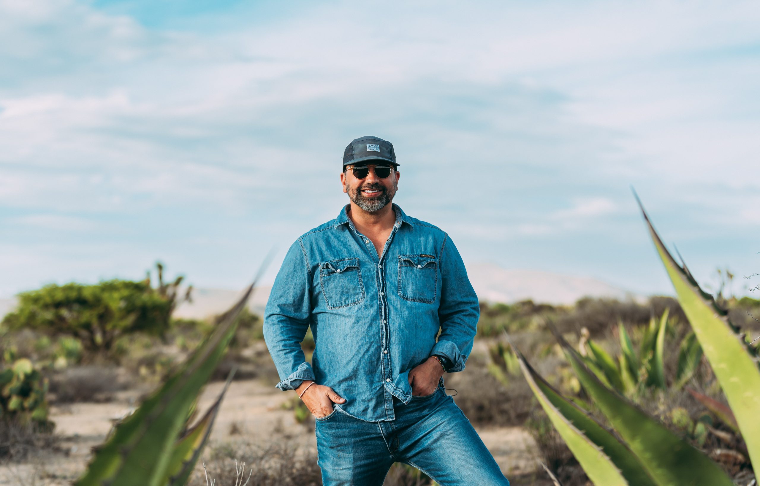 Andrew de la Torre stands among agave plants in a desert landscape, wearing a denim shirt, jeans, baseball cap and sunglasses, smiling at the camera.