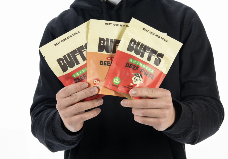 A person holds three bags of Buffs beef puffs fanned out, with the “Meat Your New Snack” tagline visible across the tops.