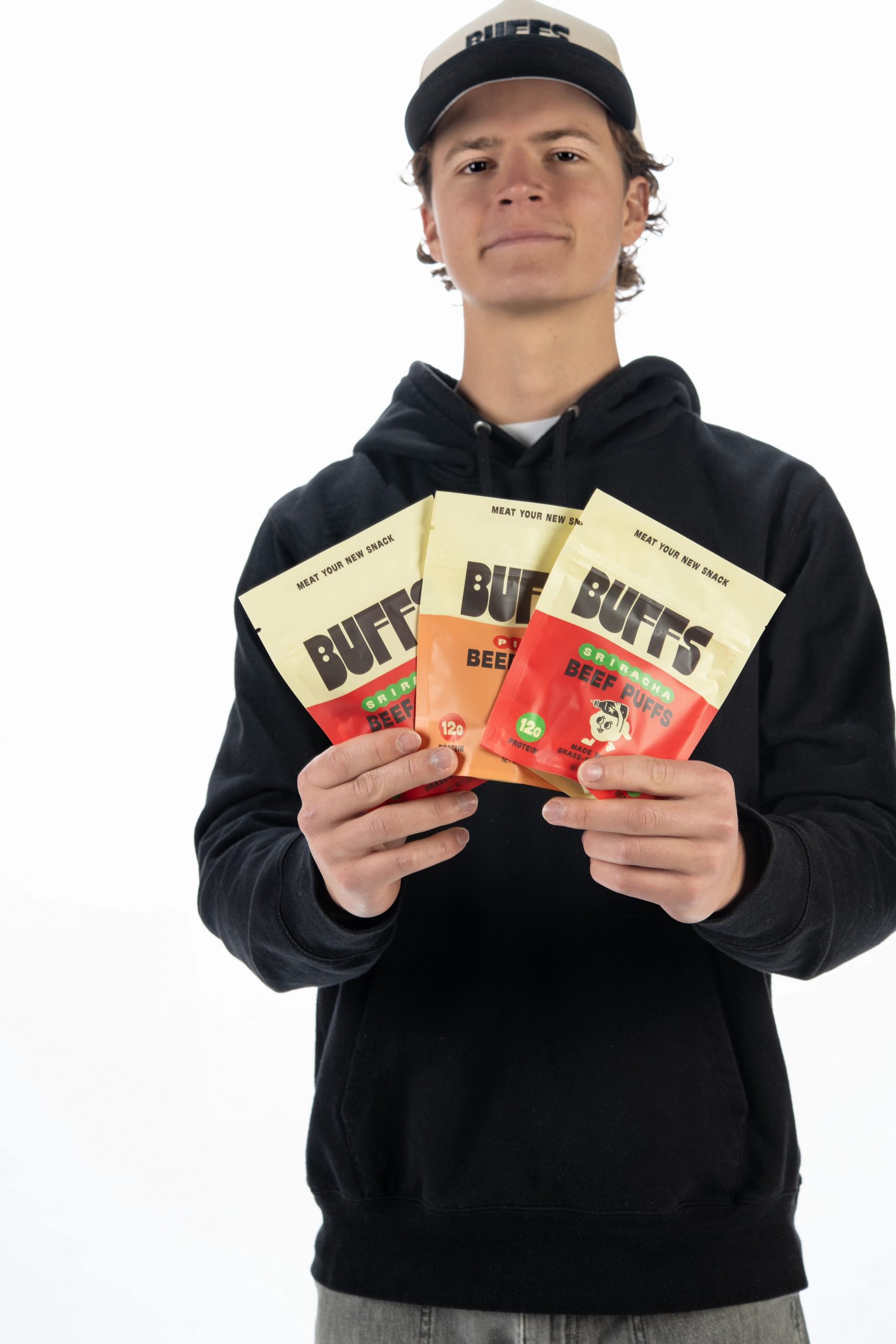 Beckett Kitaen stands against a white background, wearing a black hoodie and a light-colored cap, holding three bags of Buffs beef puffs fanned out in both hands toward the camera.