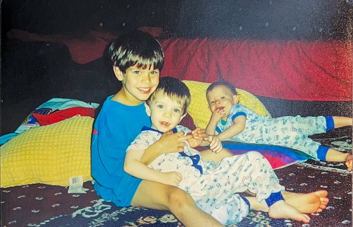 Austin, Nick and Drew Perrotti sit as children on a navy and maroon patterned carpet, smiling toward the camera.