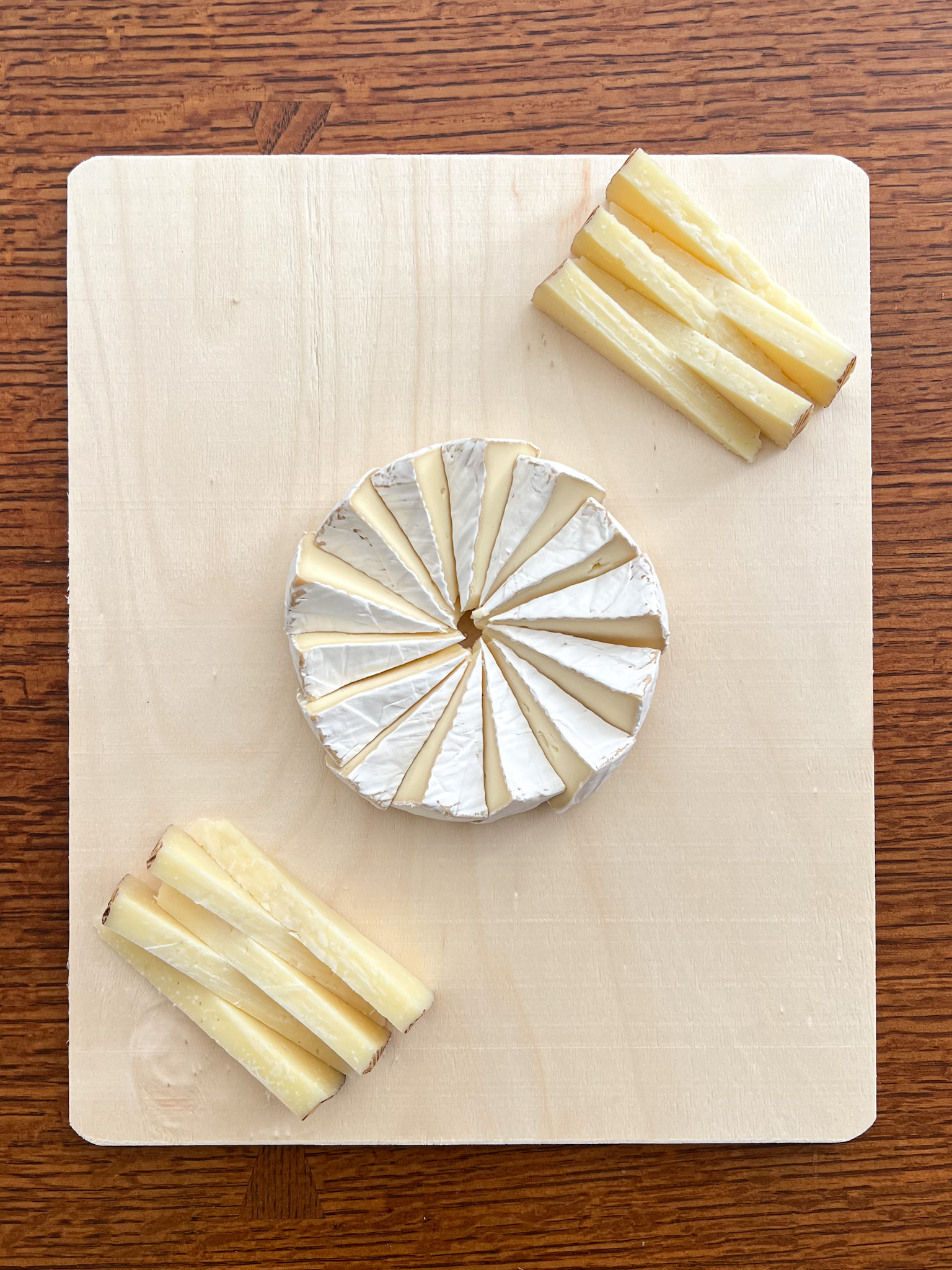A wheel of brie arranged in a pinwheel pattern at the center of a wood serving board, with two stacks of pale yellow cheese wedges placed in opposite corners.