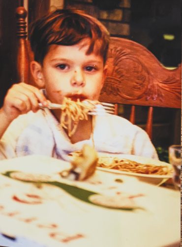 A young Austin Perrotti sits in a wooden chair eating spaghetti with a fork at a table. A white Perrotti’s Pizza box sits in the foreground.