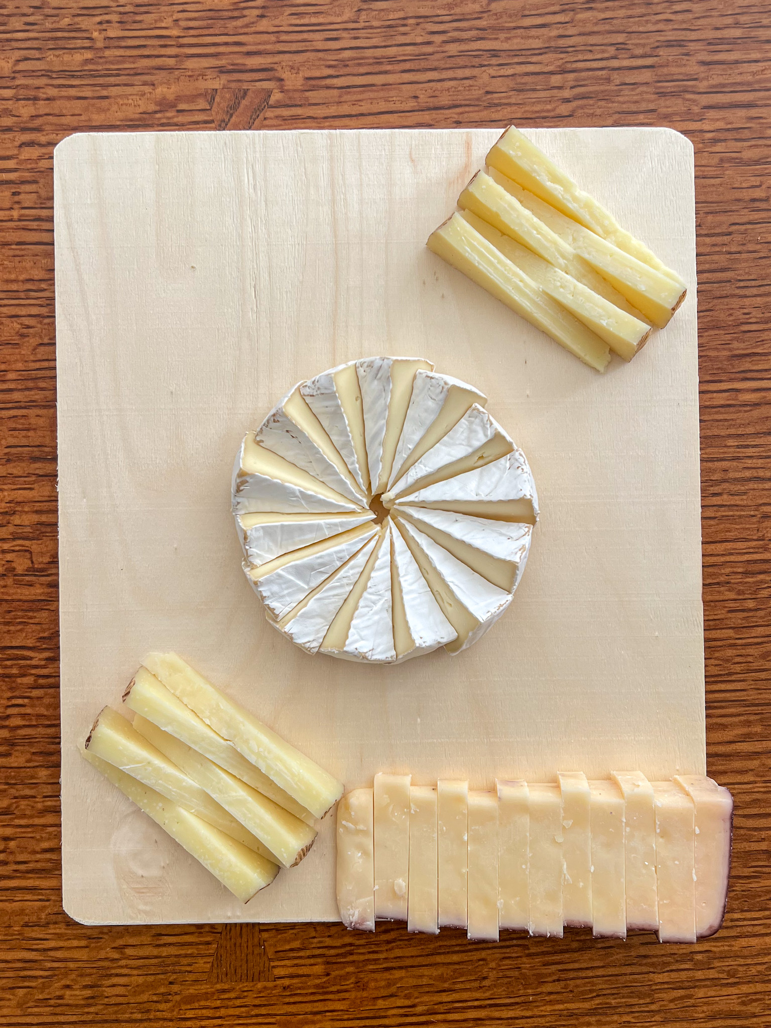 A wheel of brie arranged in a pinwheel pattern at the center of a wood serving board, with two stacks of pale yellow cheese wedges in opposite corners and a block of sliced cheese with a reddish edge along the bottom.