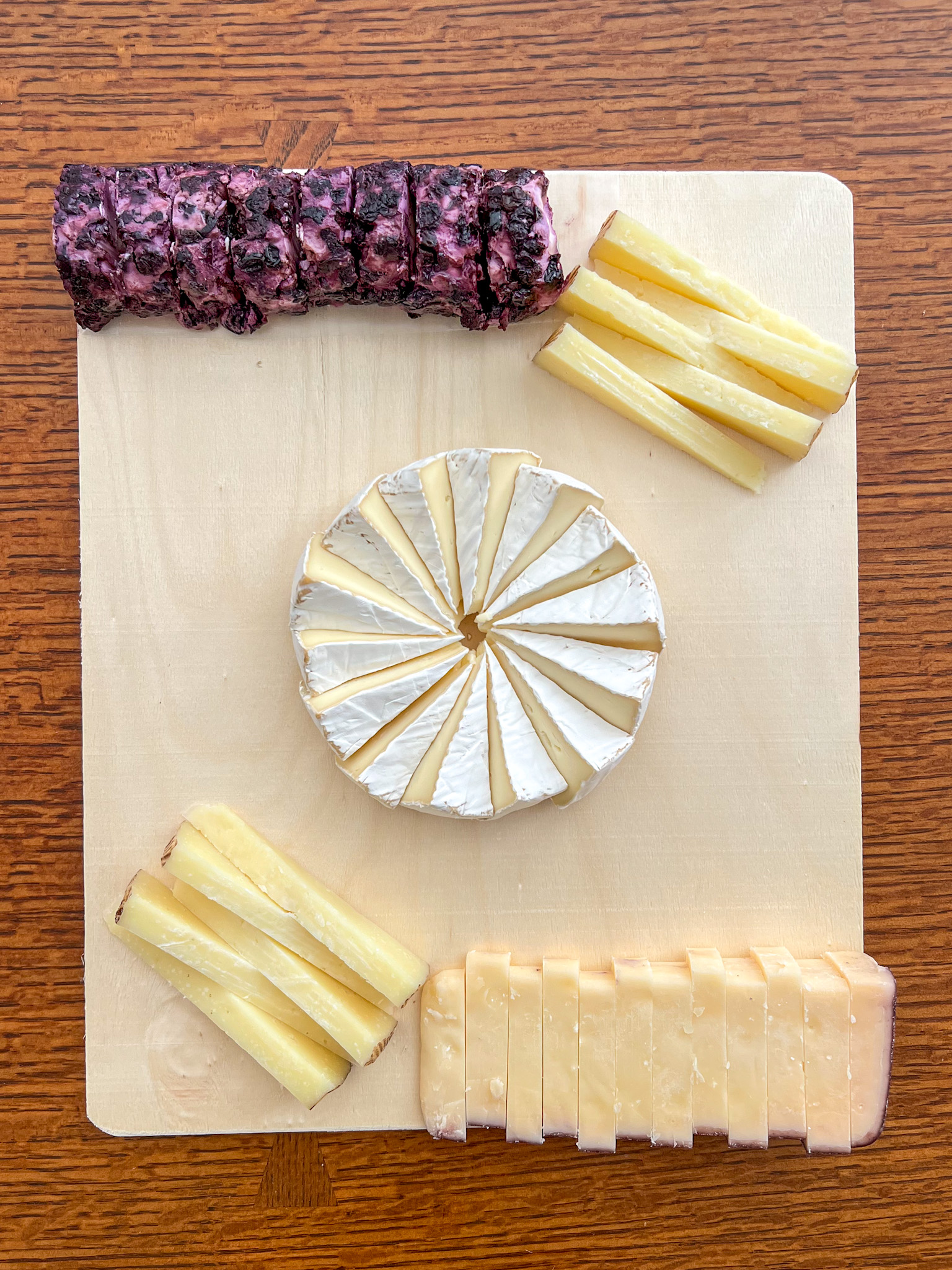 A wheel of brie arranged in a pinwheel pattern at the center of a wood serving board, with two stacks of pale yellow cheese wedges in opposite corners, a block of sliced cheese with a reddish edge along the bottom and a sliced log of purple-and-white marbled cheese along the upper edge.