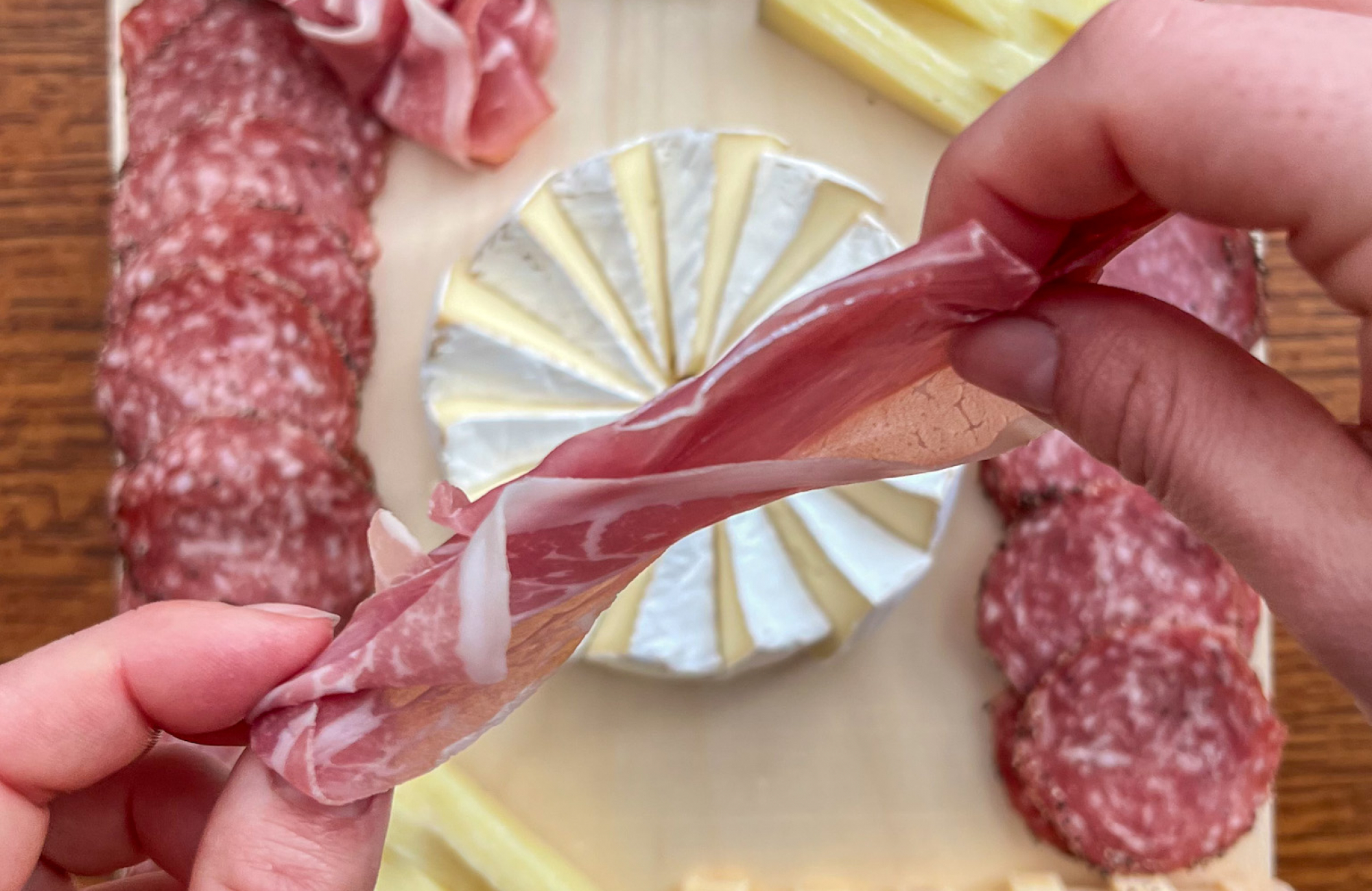 Two hands shape a slice of prosciutto above a wood serving board arranged with a brie pinwheel, rows of salami, stacks of cheese and a sliced log of purple-and-white marbled cheese.