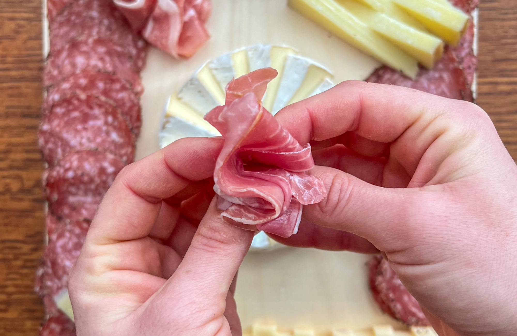 Two hands shape a slice of prosciutto above a wood serving board arranged with a brie pinwheel, rows of salami, stacks of cheese and a sliced log of purple-and-white marbled cheese.