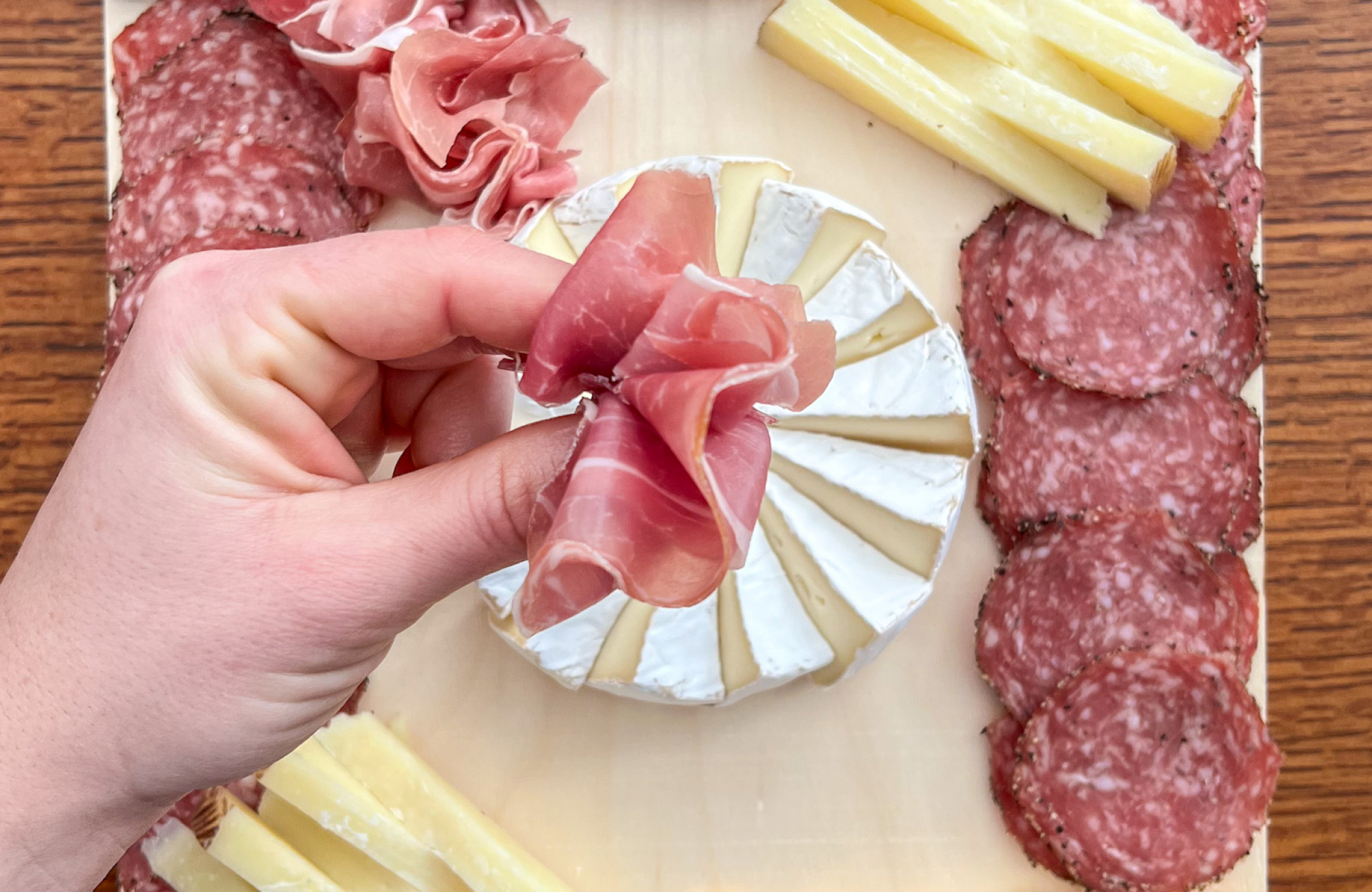 Two hands fold a slice of prosciutto into a fan shape above a wood serving board arranged with a brie pinwheel, rows of salami, stacks of cheese and a sliced log of purple-and-white marbled cheese.