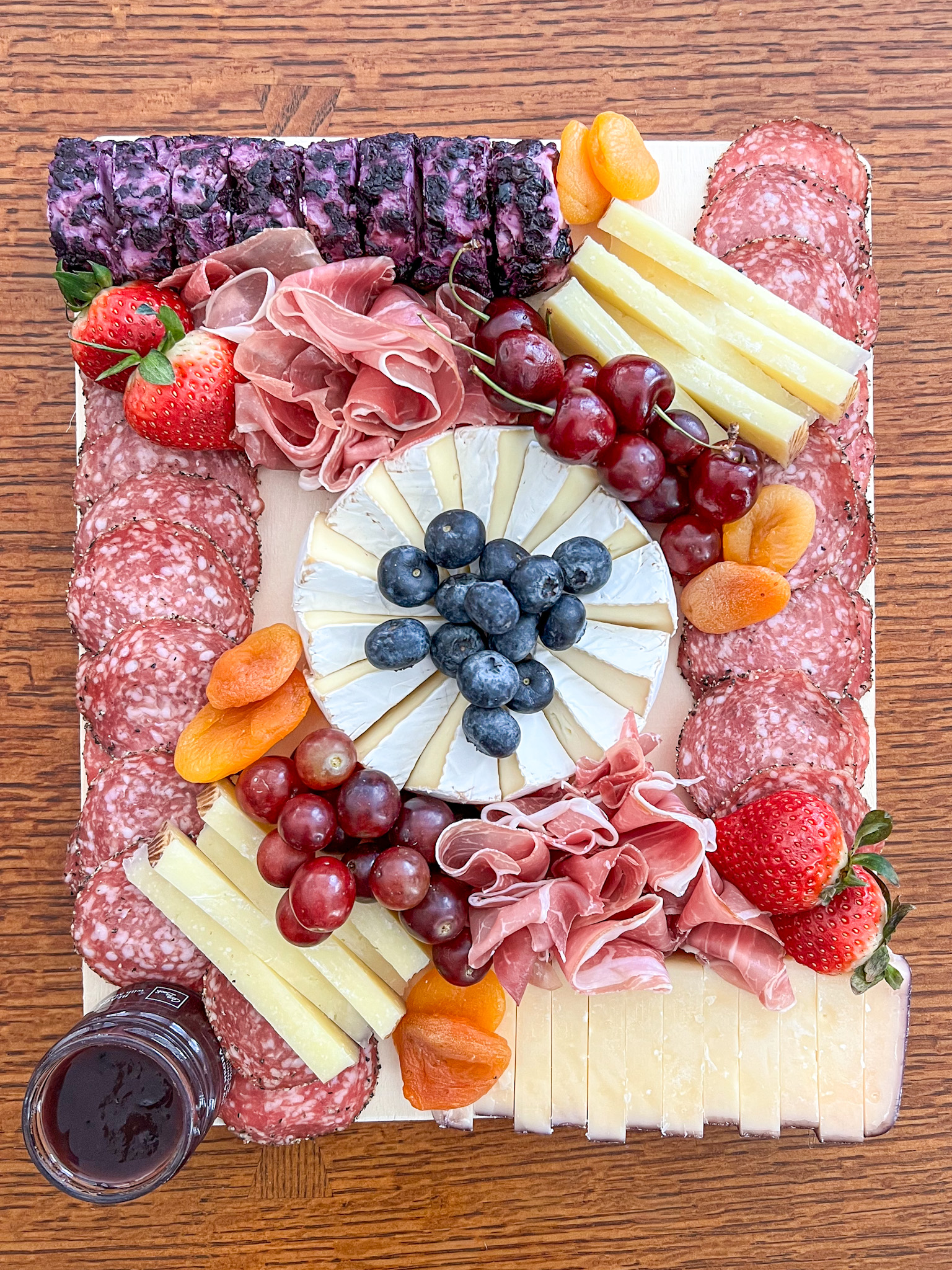 A grazing board on a wood serving platter with a wheel of brie at the center, topped with blueberries, surrounded by cherries on stems, red grapes, strawberries, dried apricots, fanned prosciutto, stacks of pale yellow cheese wedges, a sliced log of purple-and-white marbled cheese and a block of sliced cheese with a reddish edge. Rows of round sliced salami frame the long edges, with a small jar of dark spread at the corner.