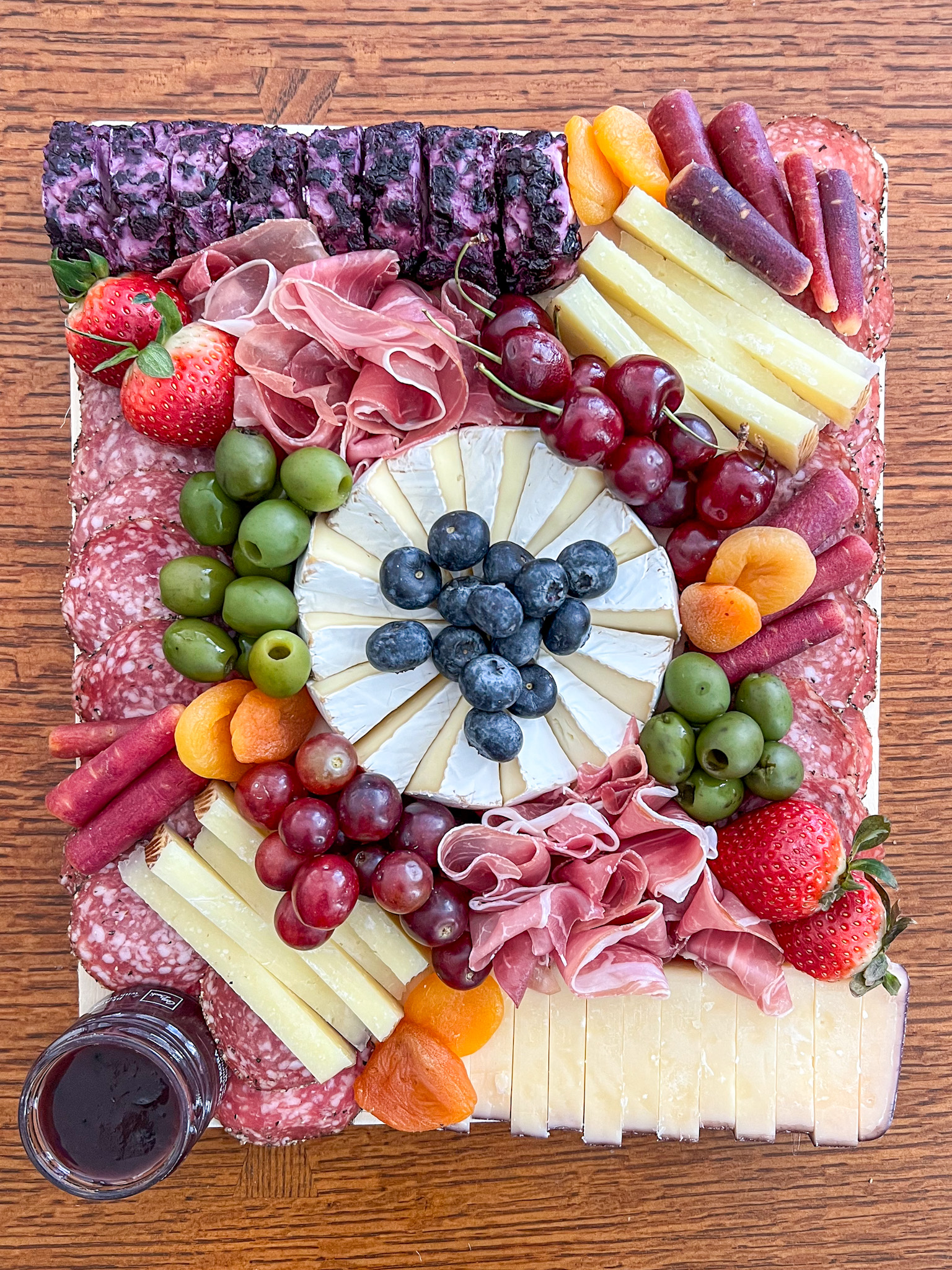 A grazing board on a wood serving platter with a wheel of brie at the center, topped with blueberries, surrounded by green olives, cherries on stems, red grapes, strawberries, dried apricots, multicolored baby carrots, fanned prosciutto, stacks of pale yellow cheese wedges, a sliced log of purple-and-white marbled cheese and a block of sliced cheese with a reddish edge. Rows of round sliced salami frame the long edges, with a small jar of dark spread at the corner.