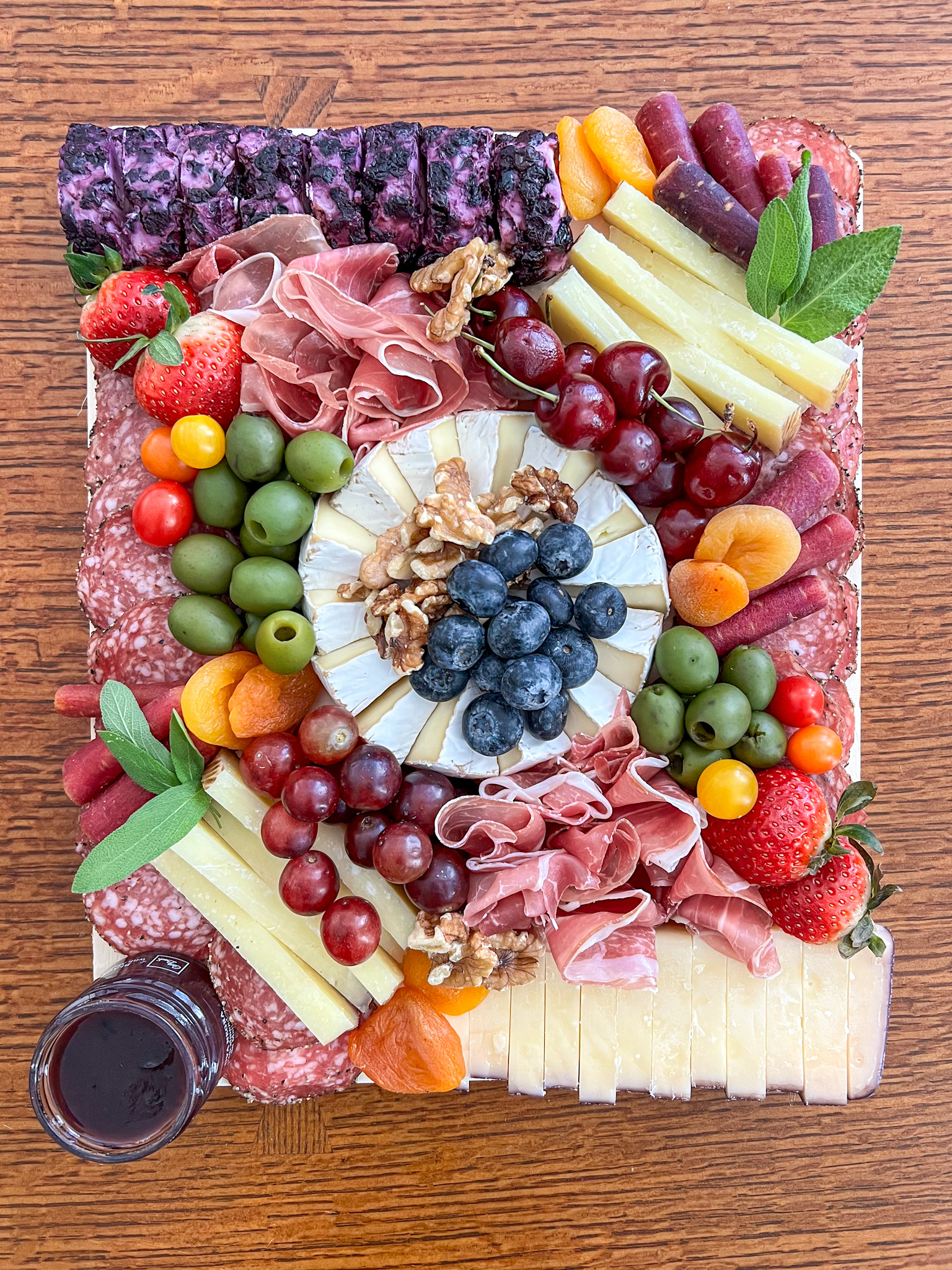 A fully assembled grazing board on a wood serving platter, with a wheel of brie at the center topped with blueberries and walnuts, surrounded by green olives, cherries on stems, red grapes, strawberries, dried apricots, multicolored baby carrots, small mixed tomatoes, fanned prosciutto, sage leaves, stacks of pale yellow cheese wedges, a sliced log of purple-and-white marbled cheese and a block of sliced cheese with a reddish edge. Rows of round sliced salami frame the long edges, with a small jar of dark spread at the corner.