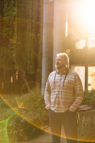 A bearded man with a long braid, wearing a striped flannel shirt, stands with his hands in his pockets and looks off to the side, backlit by warm golden sunlight with lens flare around him and a courtyard of greenery and concrete columns behind.