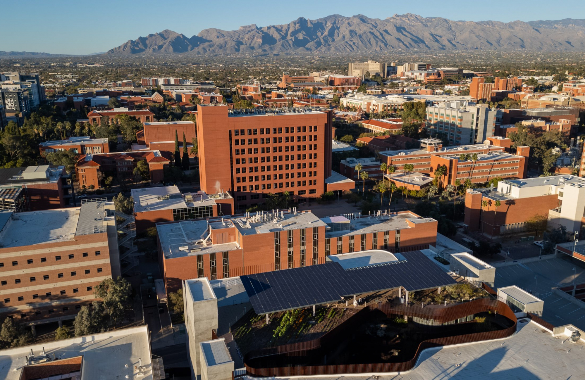 Aerial view of red-brick University of Arizona buildings with a rooftop solar array and planted beds in the foreground and mountains rising in the distance.