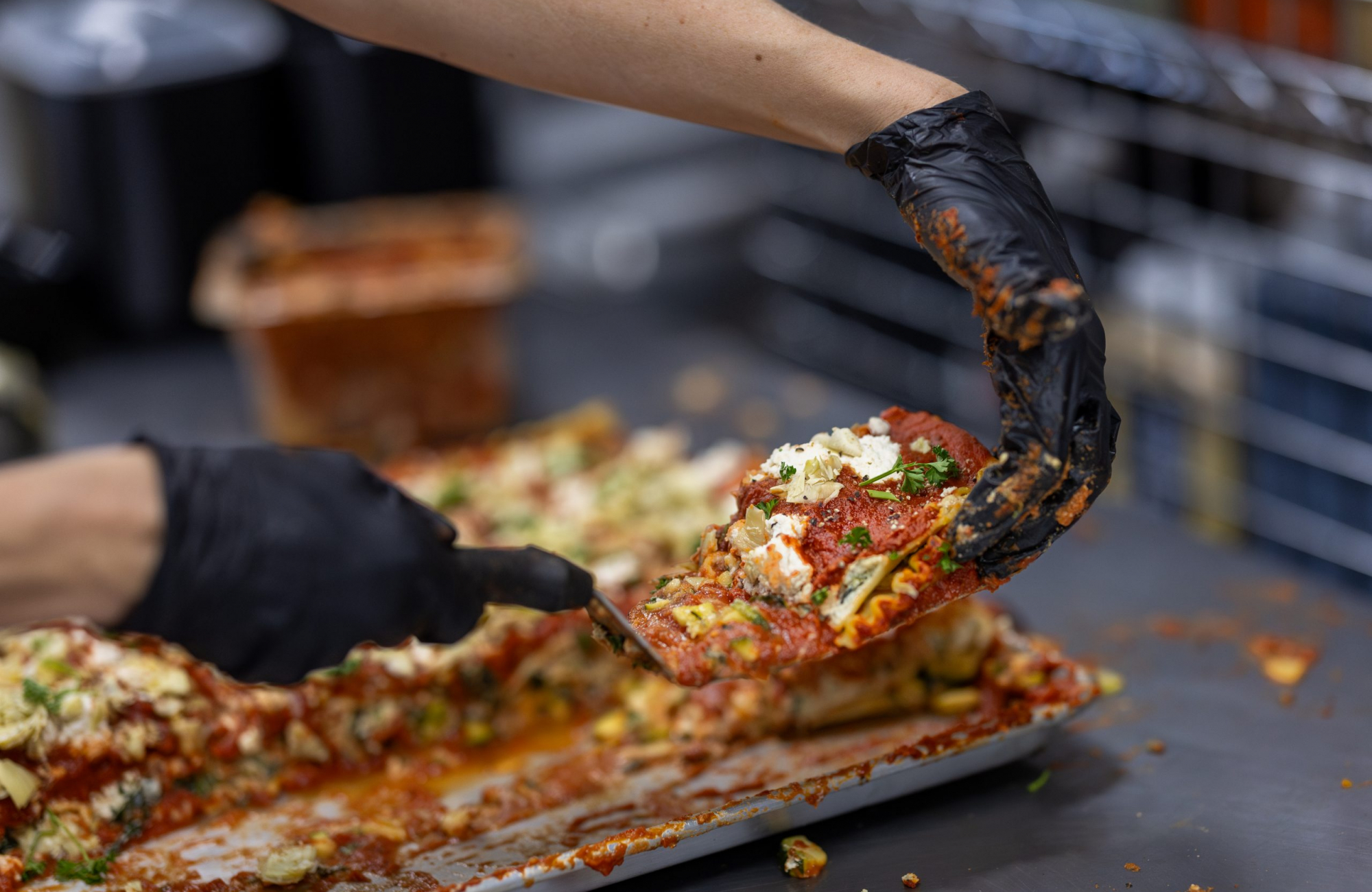 Two gloved hands lift a slice of vegan lasagna from a sheet pan, showing layers of pasta, red sauce, vegetables and plant-based cheese.