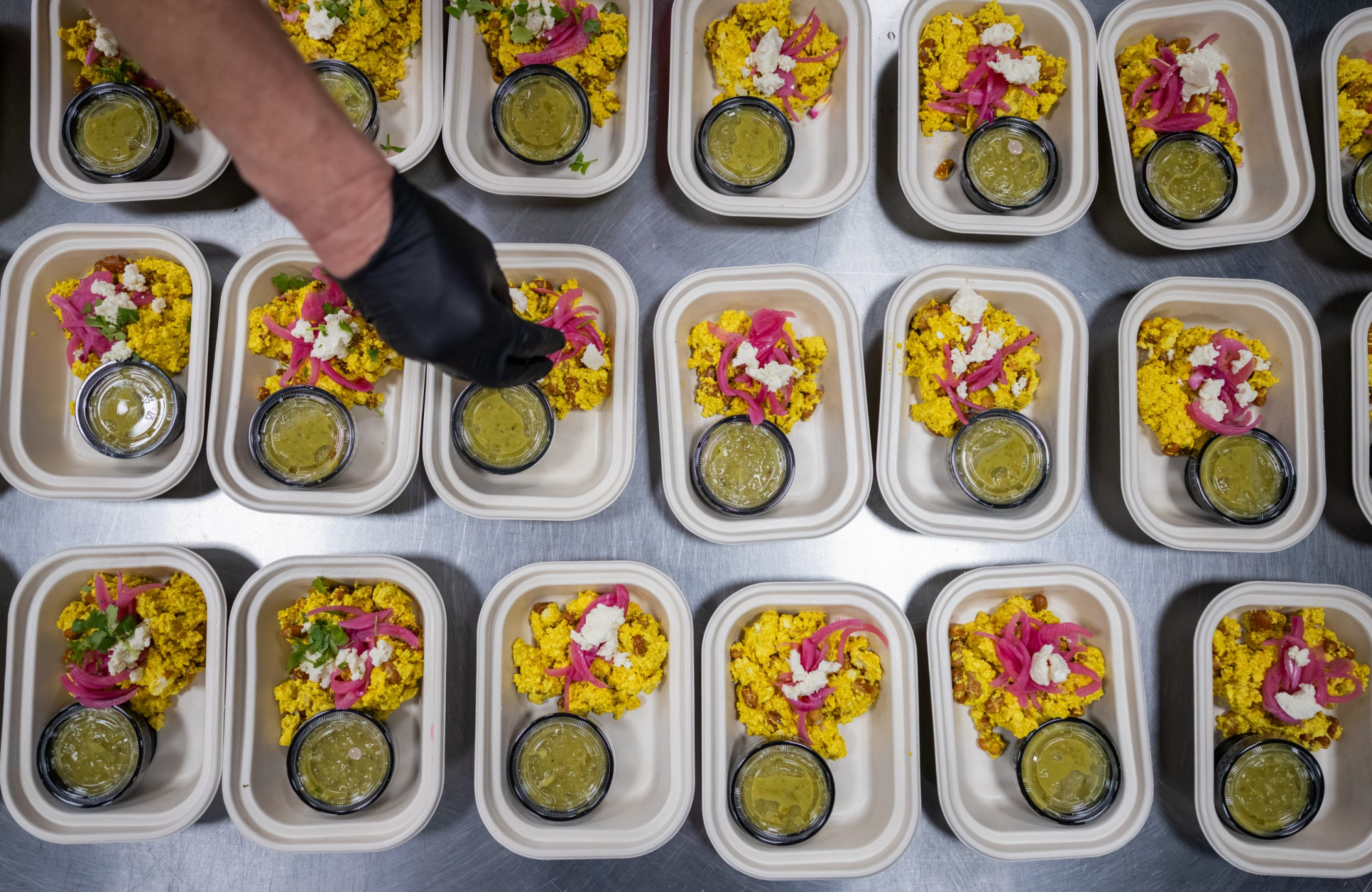 Overhead view of rows of takeout containers filled with yellow tofu scramble, pinto beans, pickled red onions and salsa verde, with a gloved hand reaching in to garnish one.