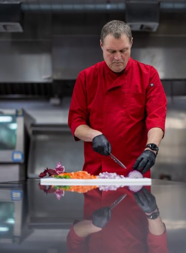 Jason Wyrick, in a red chef’s coat and black gloves, chops vegetables at a stainless steel prep table, with diced red onion, white onion, carrot, green pepper and red cabbage arrayed on a white cutting board in front of him.