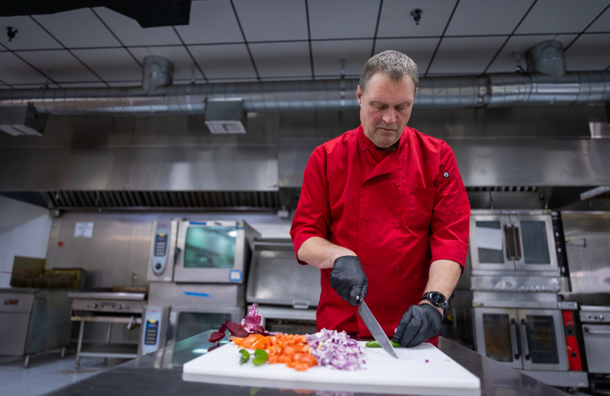 Jason Wyrick, in a red chef's coat and black gloves, chops vegetables on a white cutting board in a stainless steel commercial kitchen.