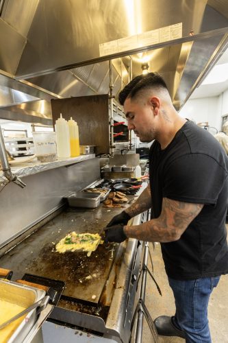 Reale works the flat-top griddle at Paris Coffee Shop, folding an omelet as sausage patties and bacon cook nearby.