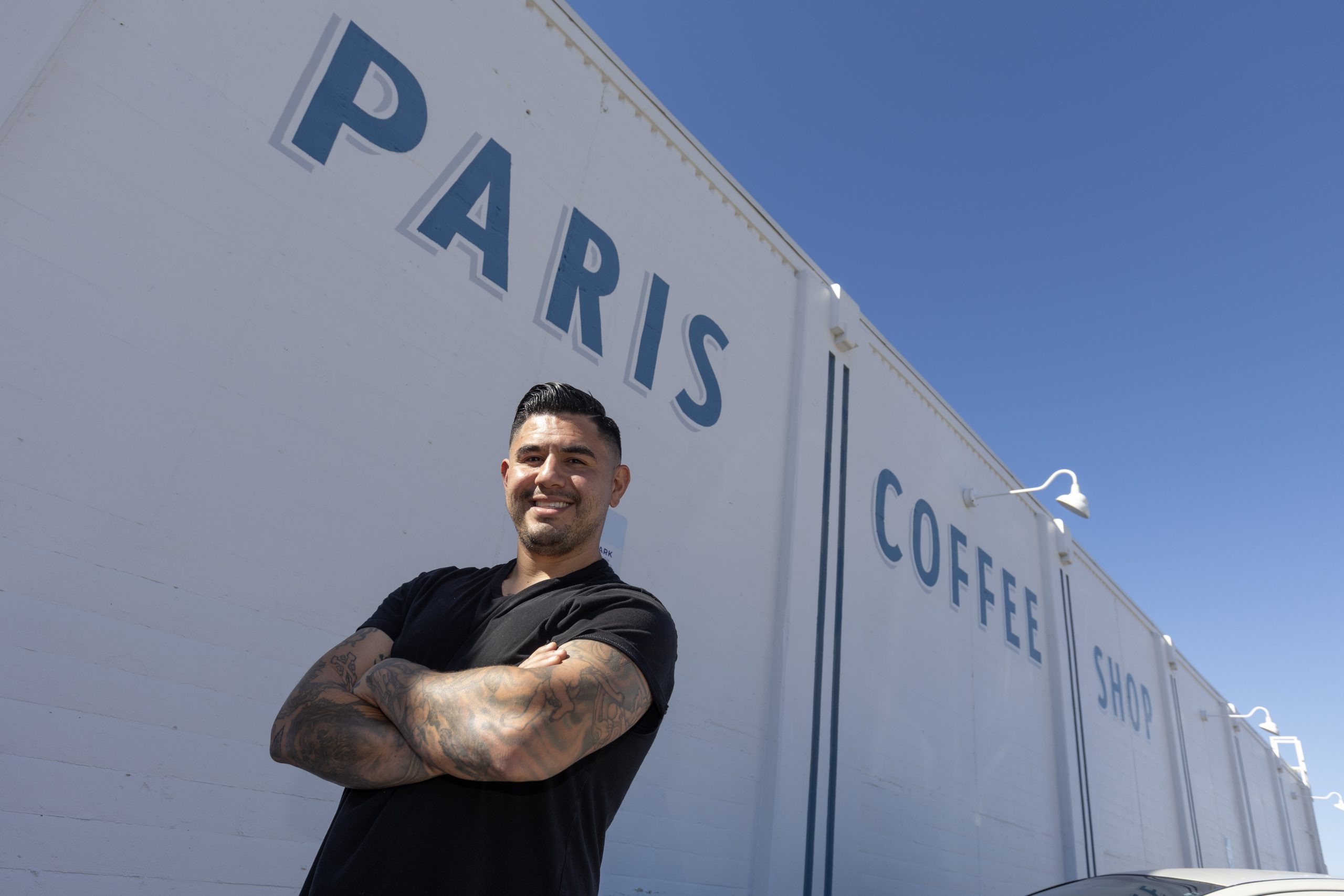 Chris Reale stands with arms crossed outside Paris Coffee Shop, with the diner's large blue “PARIS COFFEE SHOP” lettering painted on the white exterior wall behind him.