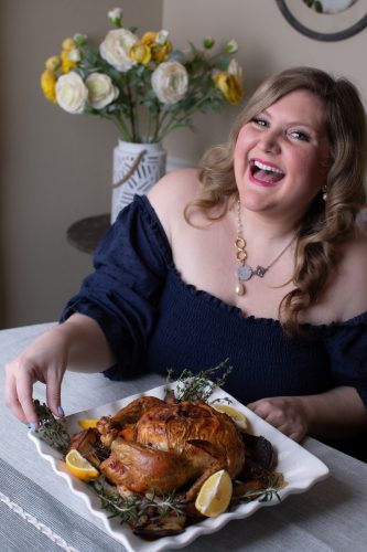 Katie-Rose Watson laughs at a table with a platter of whole roasted poultry garnished with herbs and citrus, a vase of flowers behind her.