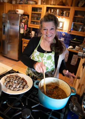 June Naylor Harris smiles at the camera while stirring a teal Dutch oven on a gas stovetop, a plate of cubed browned meat beside her. She wears a white apron printed with green leafy produce over a black top.