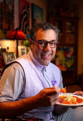 Lanny Lancarte II, in a lavender TCU vest and tortoiseshell glasses, smiles while holding a plate of enchiladas marked “J. Garcia’s.”