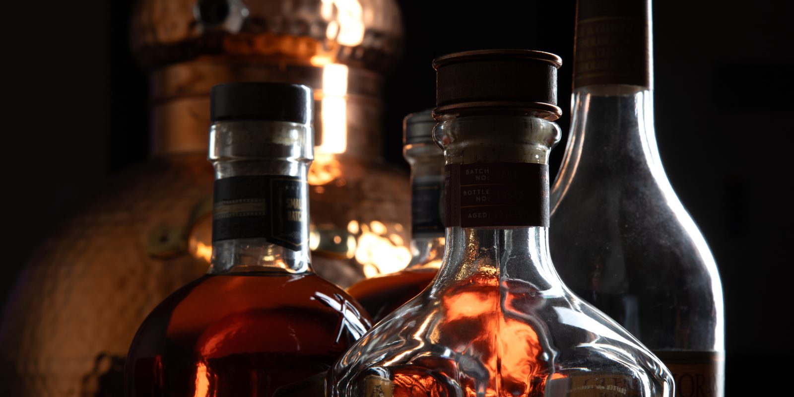Three whiskey bottles backlit in warm amber light, with a copper pot still visible in the dark background.