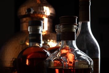 Three whiskey bottles backlit in warm amber light, with a copper pot still visible in the dark background.