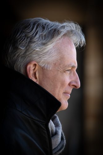 Eric Simanek photographed in profile, with silver hair and a black jacket over a gray scarf, looking off to the right against a dark background.
