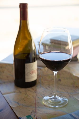 A wine bottle with a partially obscured label and a full glass of red wine sit on top of an unfolded Paris transit map, with soft, warm light filtering in from a window in the background.