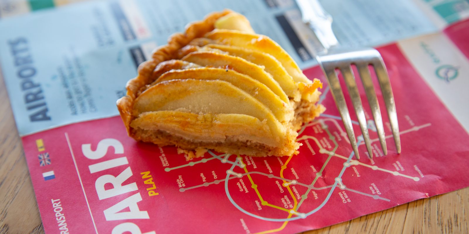 A slice of French apple tart with thinly sliced, glazed apples fanned across a flaky pastry crust, served on top of a red Paris transit map next to a silver fork on a wooden table.