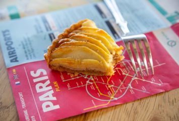 A slice of French apple tart with thinly sliced, glazed apples fanned across a flaky pastry crust, served on top of a red Paris transit map next to a silver fork on a wooden table.
