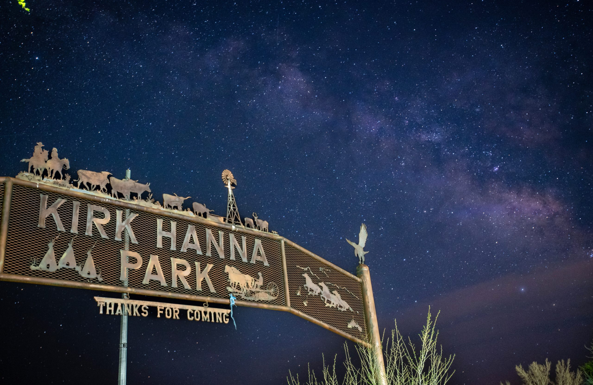 A metal archway sign reading “Kirk Hanna Park” with “Thanks for Coming” hanging below, topped with cut-metal silhouettes of cattle, horseback riders and a windmill, set against the Milky Way and a star-filled night sky.