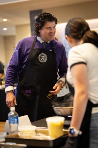 TCU Chancellor Daniel W. Pullin, wearing a purple TCU sweatshirt and apron, smiles in conversation during a cooking class with Chancellor's Scholars.