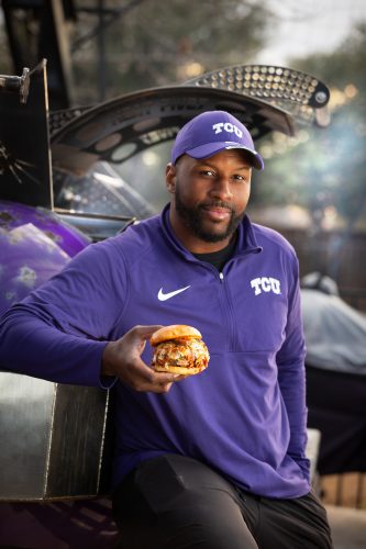 David Hawthorne, in a purple TCU quarter-zip and cap, holds a pulled pork sandwich in front of a smoker.