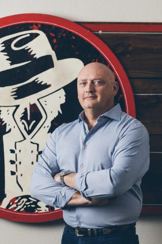 Tom Gordon stands with arms crossed in front of a large circular Slim Chickens logo featuring the brand’s cowboy mascot illustration in cream and black on a red background.