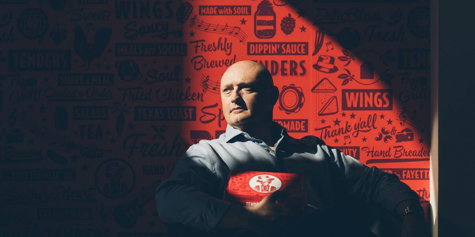 Tom Gordon, co-founder and CEO of Slim Chickens, holds a branded takeout container while seated against a red wall covered in the restaurant's signature illustrated logos and food-themed phrases.