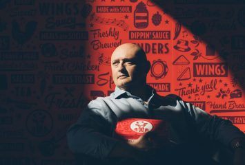 Tom Gordon, co-founder and CEO of Slim Chickens, holds a branded takeout container while seated against a red wall covered in the restaurant's signature illustrated logos and food-themed phrases.