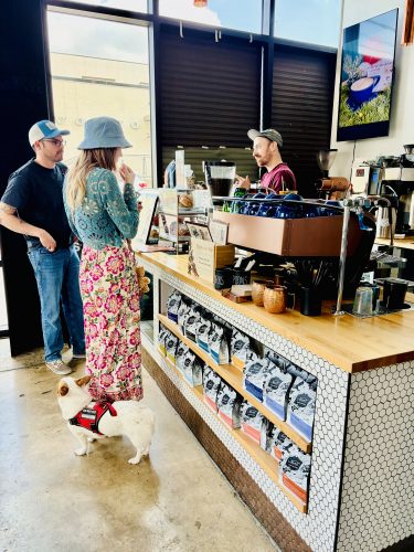 Two customers with a service dog chat with a barista at the counter of Roots Coffeehouse, which features a copper espresso machine, retail coffee bags displayed on hexagonal tile shelving, and modern industrial decor.