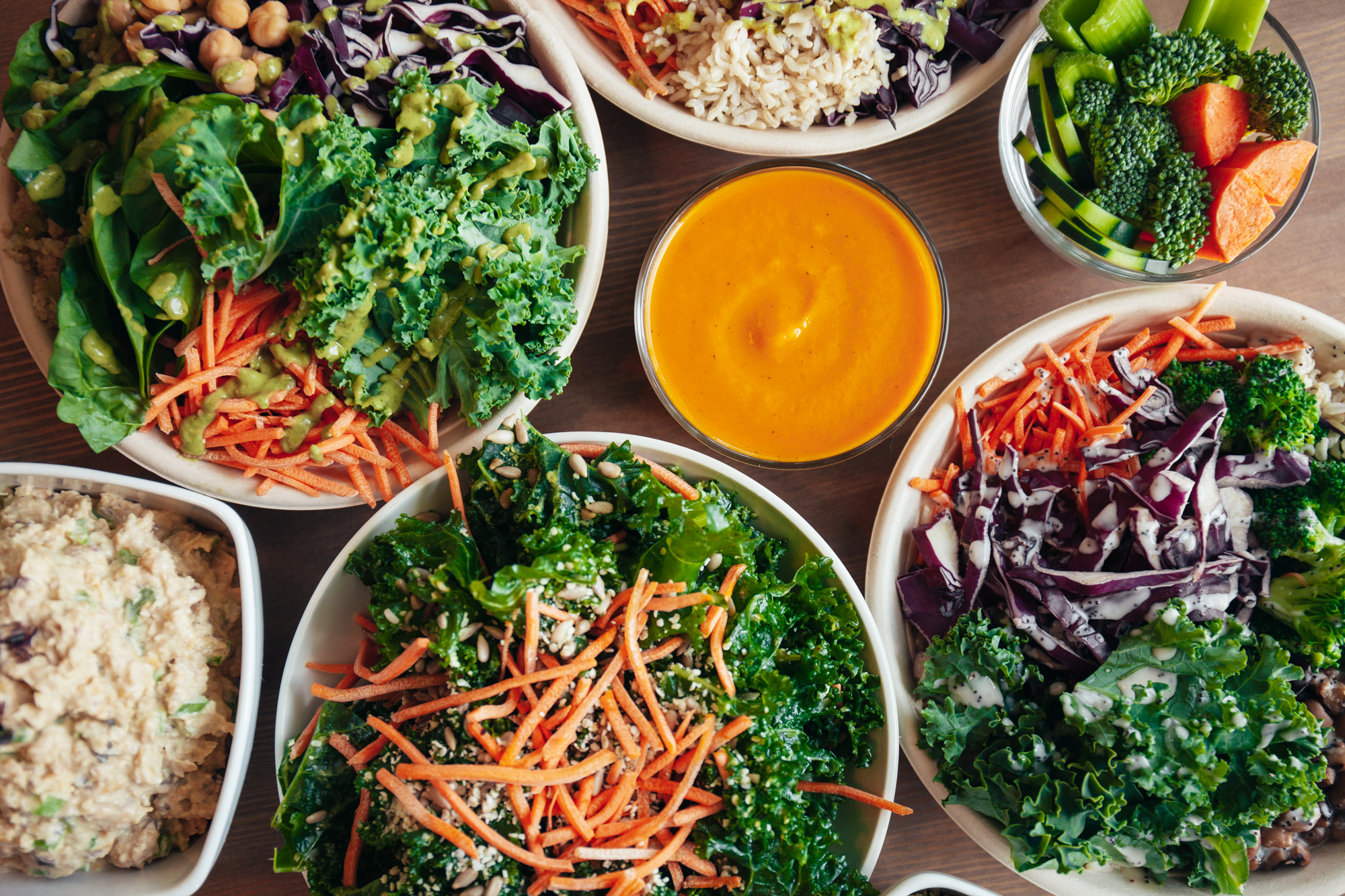 Overhead view of several colorful grain bowls arranged around a central bowl of golden-orange soup or dressing on a wooden table, featuring kale, shredded carrots, purple cabbage, broccoli, brown rice and chickpeas.