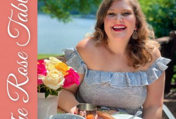 Cover of “The Rose Table” by Katie-Rose Watson, with the author smiling at an outdoor table set with a blueberry cobbler, a bowl of blueberries, whipped cream and a bouquet of pink, yellow and white roses, with a lake in the background.