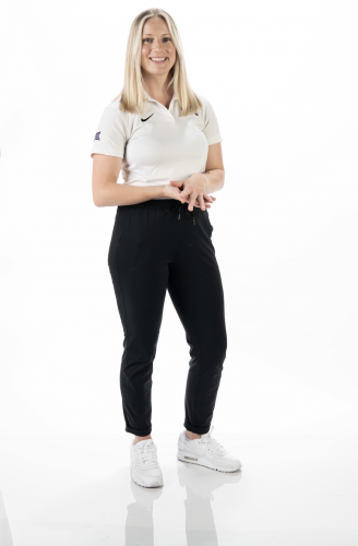 Studio portrait of Jessica Hylander, TCU assistant athletics director for sports nutrition, smiling with hands clasped, wearing a white Nike polo with TCU and Big 12 logos, black pants and white sneakers.