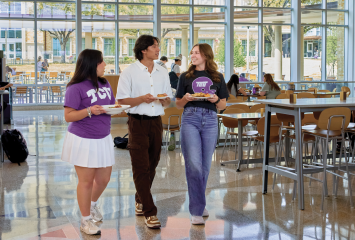 Three TCU students carry plates of food through Gutierrez Hall's sunlit dining commons.