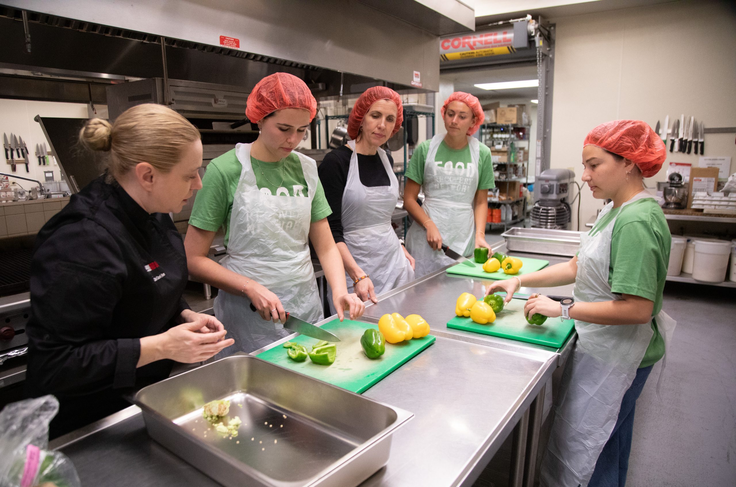 Four volunteers in green Food Recovery Network T-shirts, white aprons and red hairnets chop yellow and green bell peppers on cutting boards at a stainless-steel counter while a chef in a black uniform looks on in an industrial kitchen.