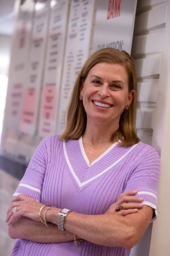 Julie Butner, TCU class of 1988 alumna and president and CEO of Tarrant Area Food Bank, stands with arms crossed in a lavender knit top, smiling in front of a wall-mounted display showing the food bank’s mission statement.