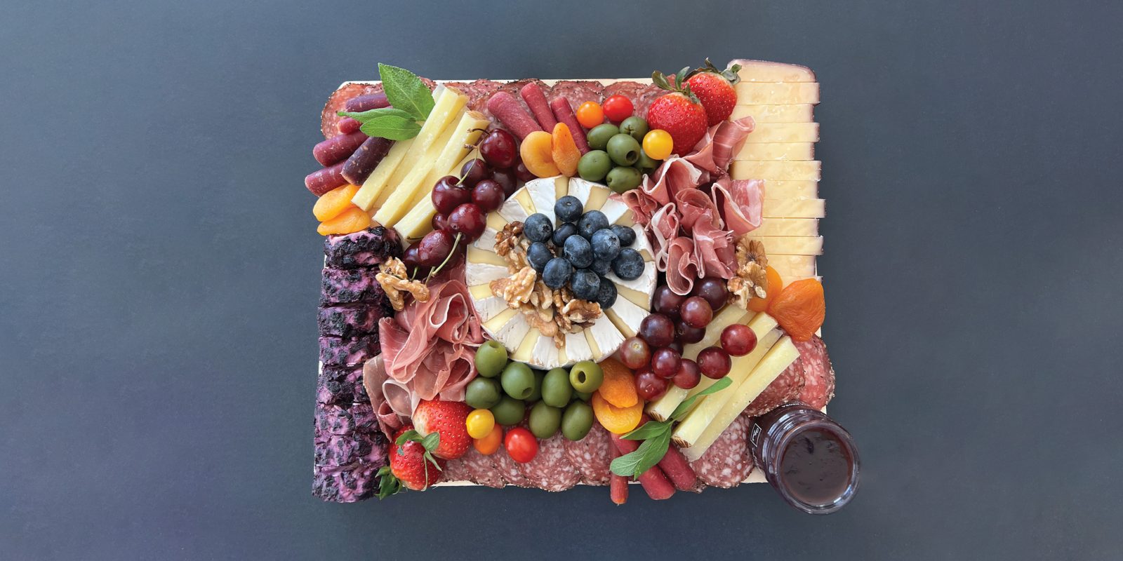 A grazing board photographed from above, with a wheel of brie at the center topped with blueberries and walnuts, ringed by folded prosciutto, sliced salami, manchego, wine-infused cheese, blueberry chèvre, cherries, strawberries, grapes, green olives, purple carrots and dried apricots.