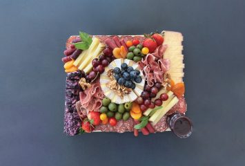 A grazing board photographed from above, with a wheel of brie at the center topped with blueberries and walnuts, ringed by folded prosciutto, sliced salami, manchego, wine-infused cheese, blueberry chèvre, cherries, strawberries, grapes, green olives, purple carrots and dried apricots.