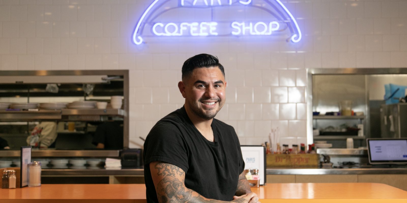 TCU alumnus Chris Reale, operating partner of Paris Coffee Shop, leans against the diner’s counter in a black T-shirt, tattoos visible on his arm, with the restaurant’s iconic blue neon sign lit up behind him.