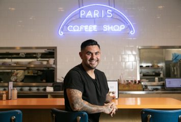 TCU alumnus Chris Reale, operating partner of Paris Coffee Shop, leans against the diner’s counter in a black T-shirt, tattoos visible on his arm, with the restaurant’s iconic blue neon sign lit up behind him.