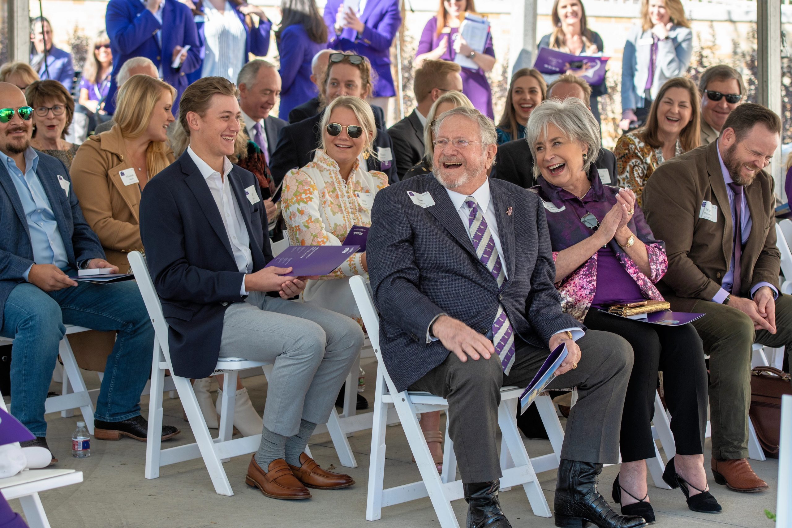 Mike Harrison laughs alongside his wife, Brenda, while seated in the front row of an outdoor TCU ceremony, surrounded by family members and a crowd of attendees in purple.