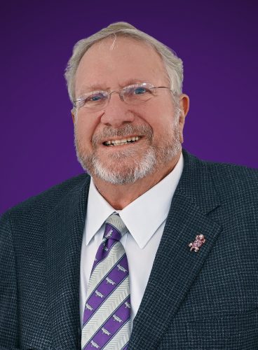 A smiling older man with a white beard and rimless glasses, wearing a charcoal blazer, white dress shirt, and purple-and-silver striped tie patterned with horned frogs, with a matching horned frog lapel pin, photographed against a solid purple background.